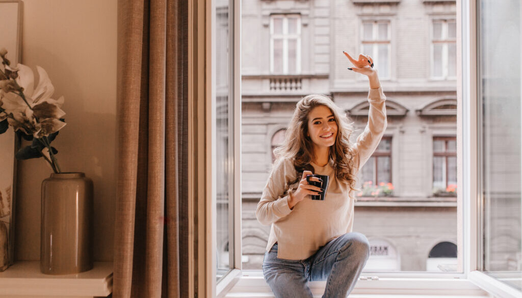 Indoor portrait of graceful white woman posing with pleasure on sill. Photo of fashionable caucasian girl sitting beside window with cup of cappuccino and enjoying day..
