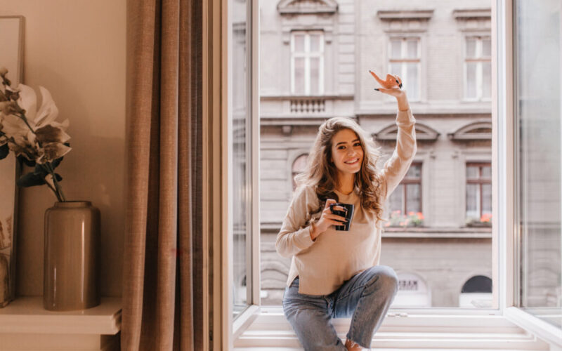 Indoor portrait of graceful white woman posing with pleasure on sill. Photo of fashionable caucasian girl sitting beside window with cup of cappuccino and enjoying day..